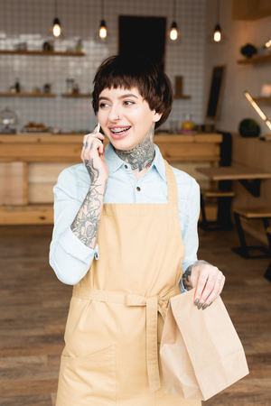 smiling waitress in apron talking on smartphone and holding paper bagの写真素材