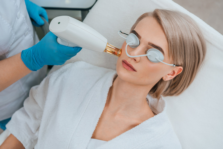 Blonde girl in protective goggles receiving laser treatment in clinicの写真素材