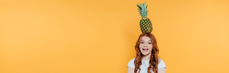 panoramic shot of beautiful excited redhead girl posing with pineapple isolated on yellow with copy spaceの写真素材