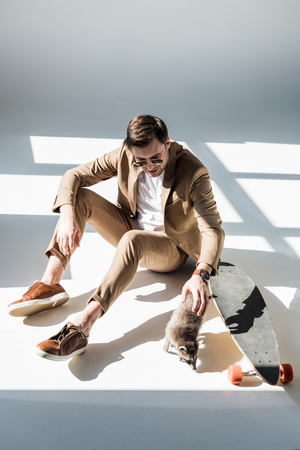 Handsome man with adorable raccoon sitting in sunlight near longboardの写真素材