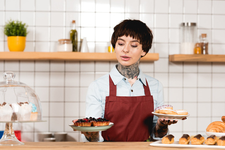 Attractive barista holding plates with delicious pastry in coffee shopの写真素材