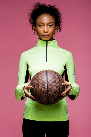 Beautiful African American sportswoman looking at camera and holding basketball isolated on pink backgroundの写真素材