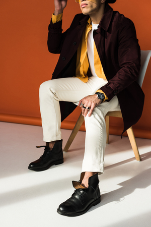 Cropped view of stylish mixed race man sitting and posing on orange backgroundの写真素材