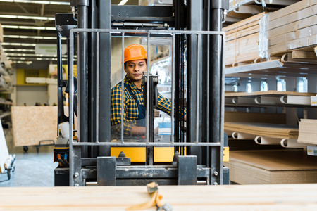 Selective focus of handsome, concentrated Indian worker sitting in forklift machine in warehouse