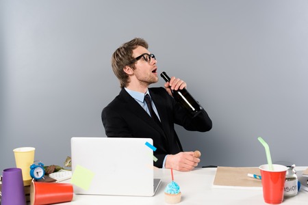 Businessman sitting at workplace with cupcake and drinking champagne from bottle isolated on grey...