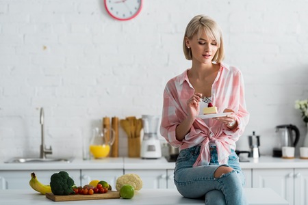 Blonde girl sitting near organic fruits and vegetables and looking at cakeの写真素材