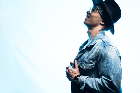 Side view of handsome mixed race man in denim and hat posing on blue with copy spaceの写真素材