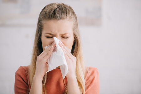 Close up of blonde woman sneezing in tissue at homeの写真素材