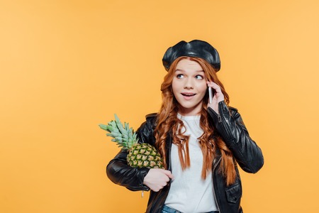 Redhead girl in leather jacket holding pineapple and talking on smartphone isolated on yellow with copy spaceの写真素材