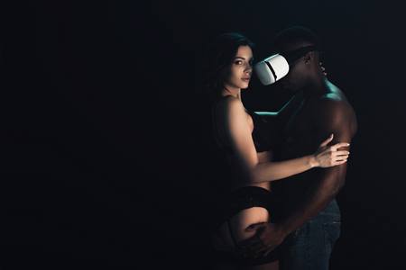 African American man in virtual reality headset embracing sexy woman in lingerie isolated on black with copy spaceの写真素材