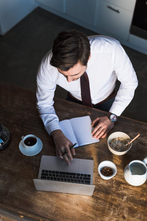 Overhead view of man using laptop and writing in notebook while having breakfast at homeの写真素材