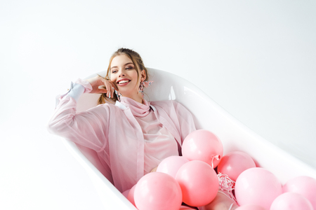 Happy young woman lying in bathtub with pink air balloons on white backgroundの写真素材