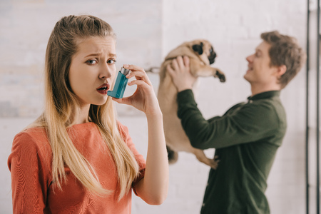 Selective focus of upset woman allergic to dog holding inhaler and looking at camera near man with pugの写真素材