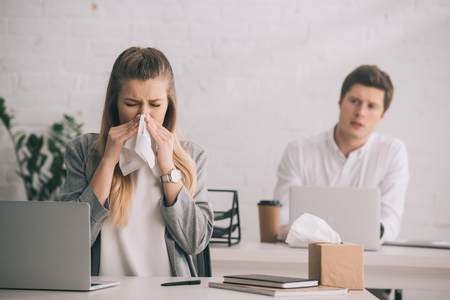 Blonde businesswoman sneezing in tissue near coworker in officeの写真素材