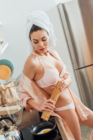 Sexy young woman in lingerie with towel on head cooking fried egg in kitchenの写真素材