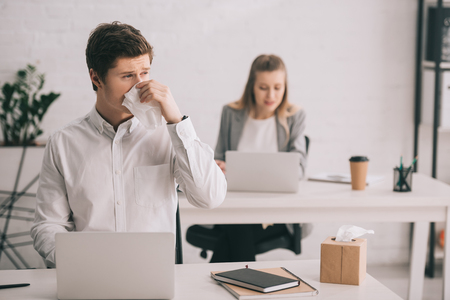 Selective focus of businessman sneezing in tissue while using laptop near businesswoman in officeの写真素材