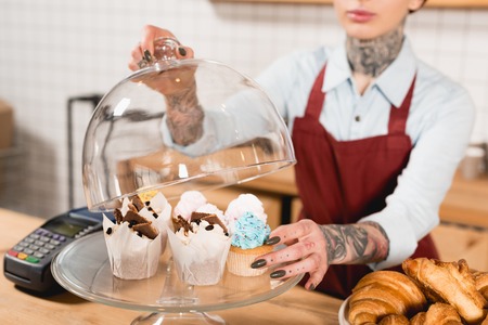 Partial view of barista in apron near bar counter with delicious desserts and payment terminal