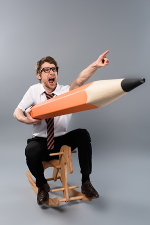 Funny businessman in glasses holding huge decorative pencil,pointing with finger, screaming and riding wooden rocking horse on grey backgroundの写真素材