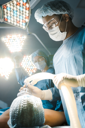 Selective focus of surgeon in uniform and goggles putting mask on patientの写真素材
