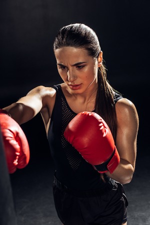 High angle view of tired boxer in red boxing gloves training with punching bag on black background
