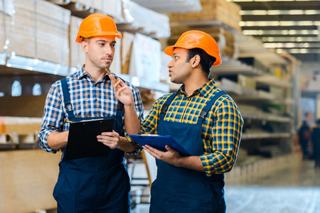 Two serious multicultural warehouse workers talking and holding clipboards