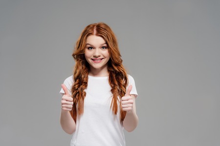 Beautiful redhead girl looking at camera, smiling and showing thumbs up isolated on grey with copy spaceの写真素材