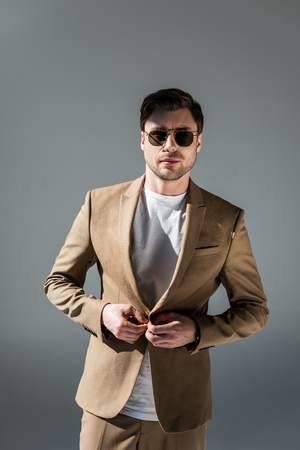 Handsome trendy man in beige suite and sunglasses looking at camera on grey backgroundの写真素材