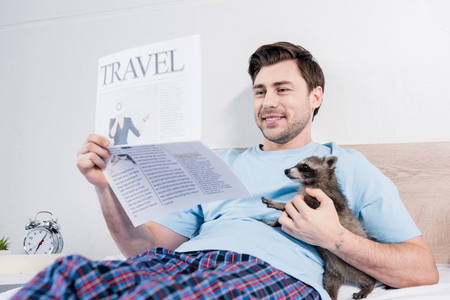 Smiling man reading travel newspaper while cuddling with funny raccoon in bedroomの写真素材