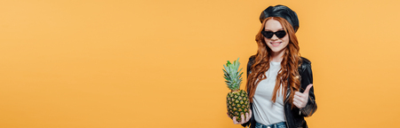 Panoramic shot of redhead girl in leather jacket with pineapple smiling and showing thumb up sign isolated on yellow backgroundの写真素材
