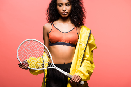 Beautiful African american sportswoman looking at camera and holding tennis racket isolated on coral color backgroundの写真素材