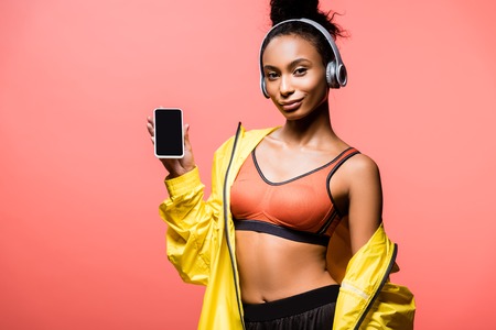 Beautiful African american sportswoman in headphones looking at camera and presenting smartphone with blank screen isolated on coral color backgroundの写真素材