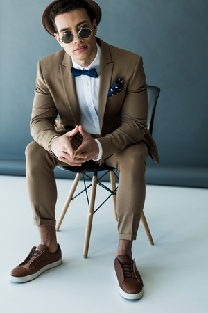 Stylish mixed race man in suit and sunglasses sitting on chair and looking at camera on grey and white backgroundの写真素材