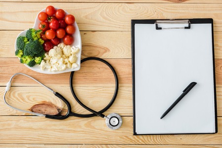 Top view of blank clipboard with pen near stethoscope and tasty vegetables on heart-shape plateの写真素材
