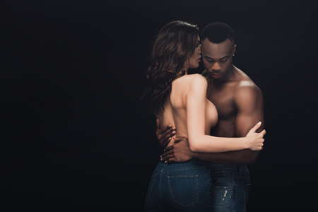 Beautiful half-naked interracial couple in denim embracing isolated on black with copy spaceの写真素材