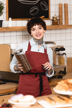 Selective focus of smiling barista holding jar with coffee grains and looking at cameraの写真素材