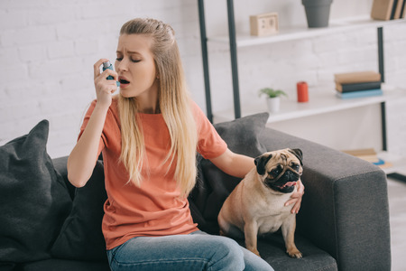 Attractive blonde woman allergic to dog using inhaler while sitting near pug at homeの写真素材