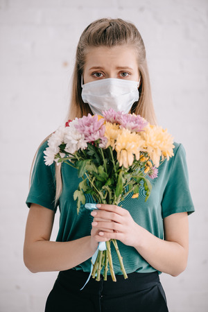 Blonde woman in medical mask holding flowers in hands and looking at cameraの写真素材