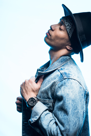 Side view of handsome stylish mixed race man in denim and hat posing isolated on blue backgroundの写真素材