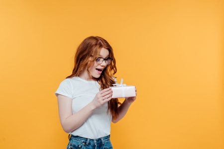 Beautiful surprised redhead girl holding gift box isolated on yellow with copy spaceの写真素材