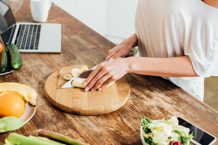 Cropped view of young woman cutting bananas with knife at table in kitchenの写真素材