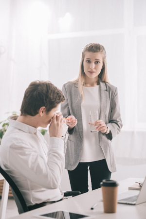 Blonde woman holding pills and glass of water near coworker sneezing in tissueの写真素材