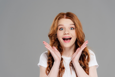 Happy redhead girl looking at camera and gesturing with hands isolated on grey backgroundの写真素材