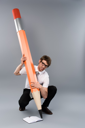 Tense businessman in glasses holding huge decorative pencil and writing in notebook on grey backgroundの写真素材