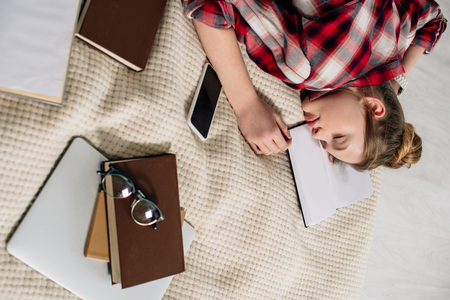 Teenager in checkered shirt sleeping on bed with books and smartphoneの写真素材