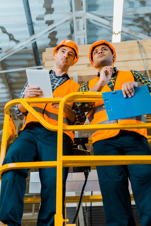 Concentrated multicultural warehouse workers with digital tablet and clipboard standing on scissor liftの写真素材