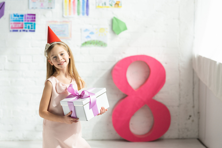 Adorable smiling kid in party cap holding present and posing near decorative pink number 8の写真素材