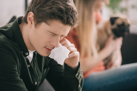 Selective focus of handsome man allergic to dog holding tissue while sitting with girl and pugの写真素材