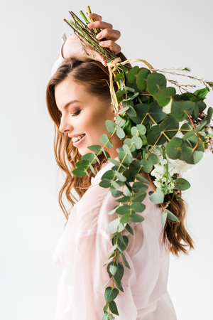 Happy beautiful girl holding flowers with green eucalyptus leaves behind back on white backgroundの写真素材