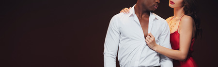 panoramic shot of woman in red dress undressing shirt of african american man isolated on black with...