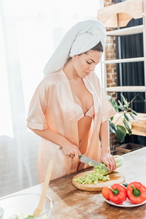 Sexy woman in housecoat with towel on head cutting celery with knife in kitchenの写真素材